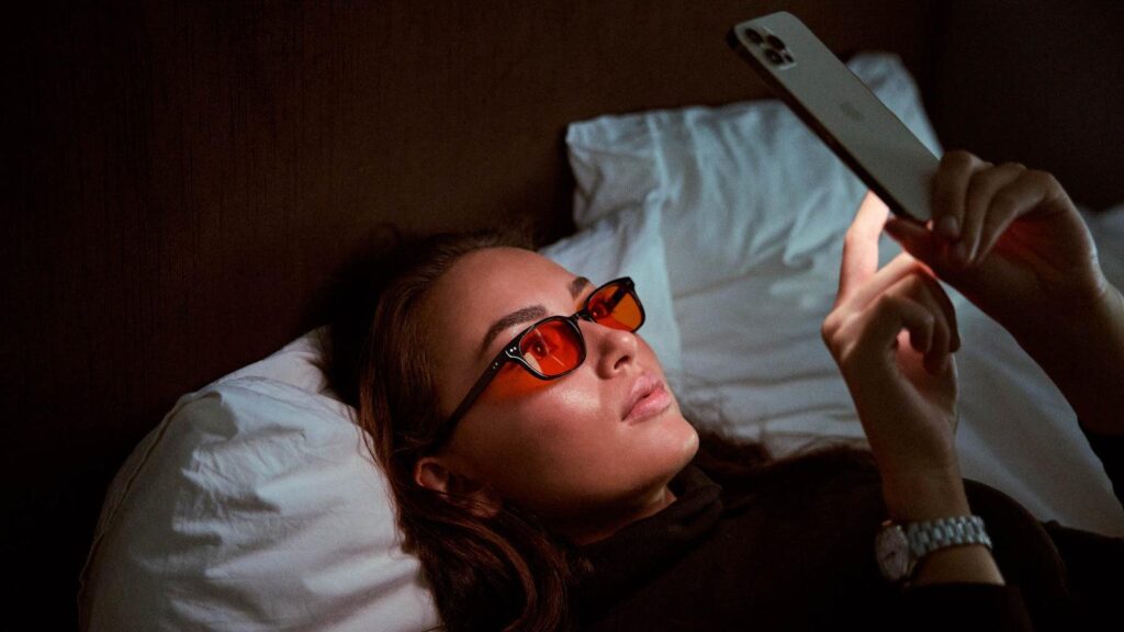 Sleep Tech: The Smart 2026 Bedroom Revolution for Deeper, Restorative Sleep. 6 Close-up of a woman in bed wearing orange-tinted sleep tech glasses while looking at a glowing phone screen.